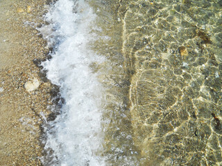 Close-up aerial view of the crystal-clear, transparent sea with gentle waves and white foam off the coast of Sardinia, Italy. The pristine Mediterranean water reveals subtle textures beneath the