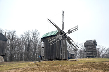Three rustic wooden windmills stand on a grassy hill against a clear, overcast sky and a backdrop of bare trees