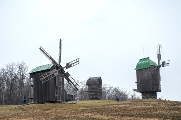 Three rustic wooden windmills stand on a grassy hill against a clear, overcast sky and a backdrop of bare trees