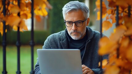 Obraz premium Middle-aged man with gray hair and glasses working on a laptop outdoors surrounded by autumn orange leaves, appearing focused and thoughtful