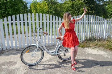 Woman wearing a red dress holding a smartphone to take a selfie next to her bicycle. The scene...