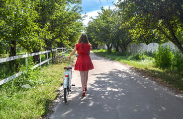 Girl in a red dress walking along a shady country road with a bicycle. The picturesque landscape is...