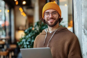 Young man wearing a mustard yellow beanie and brown hoodie smiling while holding a laptop in a cozy indoor setting with warm lighting and blurred background plants