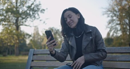 A young woman on a park bench shows a sudden shift in emotions. Initially happy and engaging with female friends on a video call, her mood turns to concern and sadness while using her phone.