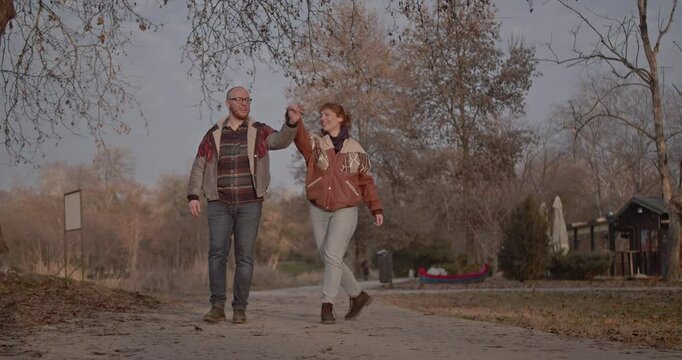 A happy couple walks hand in hand through a park, playfully swinging their arms and enjoying each other's company on a leisurely stroll.