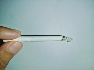 A close-up image of a person holding a burning cigarette with visible ash at the tip. 