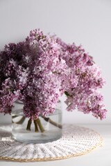 Beautiful lilac flowers in glass vase on table near white wall, closeup