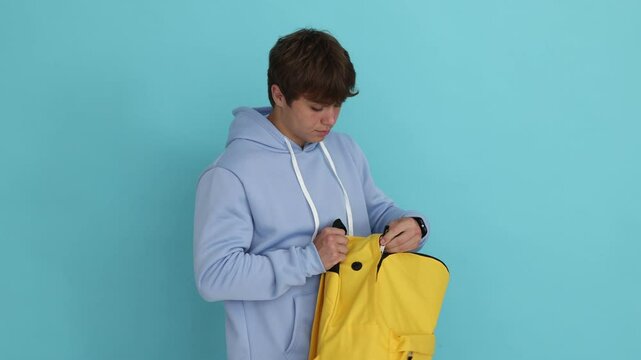 Teenage boy putting book into backpack on light blue background