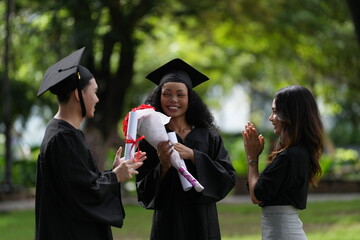 Three graduates in caps and gowns celebrating outdoors. One holds a diploma and bouquet, while the...