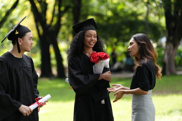 Three graduates in caps and gowns celebrating outdoors. One holds a diploma and bouquet, while the...