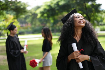 A graduate in cap and gown gazes upward with a joyful smile, holding her diploma. Behind her, two...
