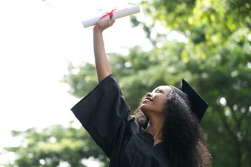 A jubilant graduate in a black cap and gown raises her diploma high in celebration, looking up with...