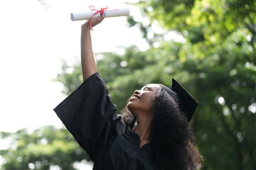 A jubilant graduate in a black cap and gown raises her diploma high in celebration, looking up with...