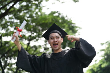 A joyful graduate in a black cap and gown proudly raises his diploma with excitement, looking...