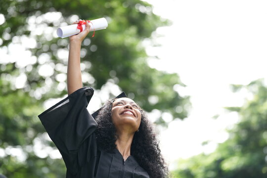 A jubilant graduate in a black cap and gown raises her diploma high in celebration, looking up with a beaming smile. The background features bright greenery, symbolizing achievement and success.