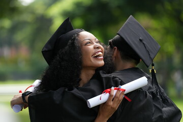 Two graduates in gowns share a warm embrace, holding diplomas with smiles of joy and pride. The...