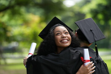 Two graduates in gowns share a warm embrace, holding diplomas with smiles of joy and pride. The...
