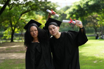 Two graduates in caps and gowns joyfully celebrate, each holding a diploma with smiles and raised...