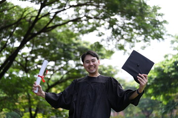 A joyful graduate in a black gown runs across a green field, holding his diploma and a folder,...