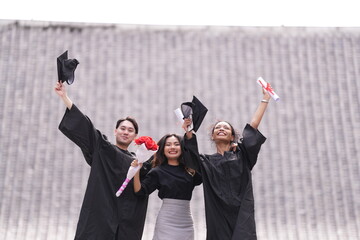 Three graduates celebrate with raised caps and diplomas, smiling joyfully. The two in gowns stand...