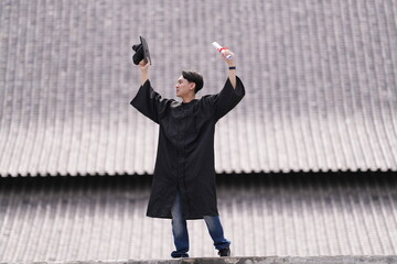 A proud graduate in a black gown holds his cap and diploma high in celebration, smiling against a...