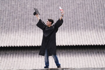 A proud graduate in a black gown holds his cap and diploma high in celebration, smiling against a...