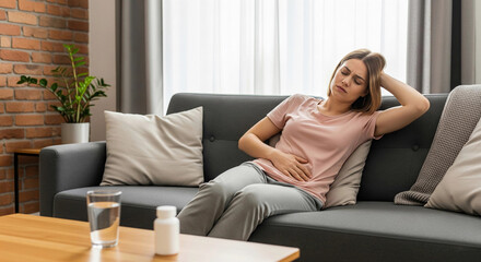 Fototapeta premium Woman experiencing abdominal pain while resting on a sofa at home, possibly due to stomach ache, menstruation, or indigestion. Ideal image for health, wellness, and medical-related content.