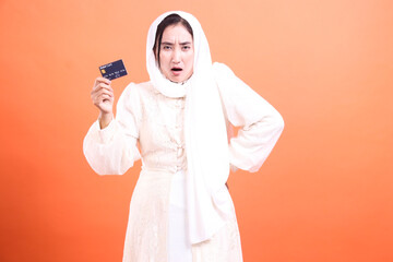 Frustrated Muslim Woman Holding Credit Card with Shocked Expression, isolated on a orange...