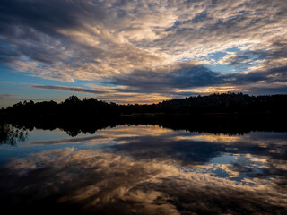 Early light as a storm approaches and clouds and sunrise are reflected on a lake with undulating forest silhouetted in the backdrop