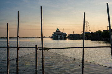 A beautiful mosque with a green dome stands over calm water, framed by fishing poles and nets at...