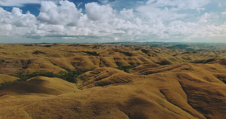 Aerial drone footage revealing the stunning Wairinding Hill landscape on Sumba Island, Indonesia, with golden rolling hills under a vibrant sky and fluffy white clouds