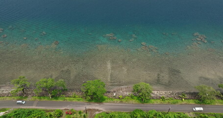 Fiji, Savu-Savu island: Aerial view of transparent shallow waters with rocks and corals alongside a coastal road, car driving countryside, lush vegetation, sunny day light. Aerial view drone footage