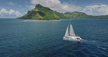 White catamaran sailing near a tropical island in French Polynesia, with lush green vegetation and...