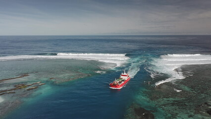 Red cargo ship is sailing in the ocean near a coral reef with breaking waves, creating a captivating scene of maritime activity and natural beauty