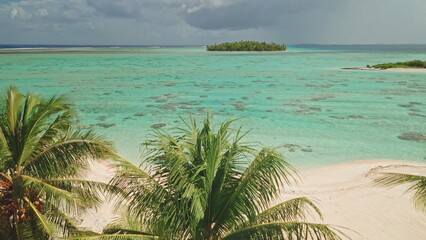Overhead view captures the stunning turquoise waters surrounding a remote island, flanked by lush palm trees and sandy beaches under a dramatic sky with distant clouds.