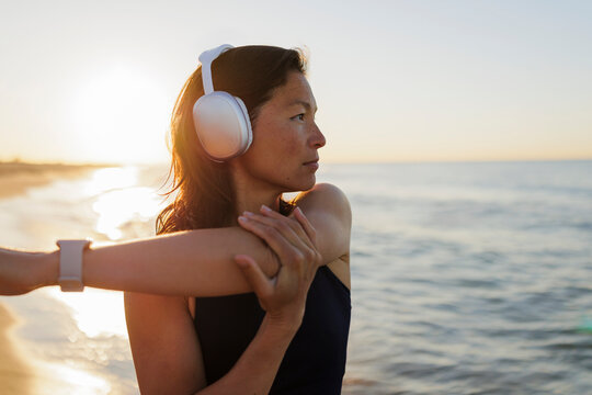 Fit woman wearing headphones stretching arms at the beach