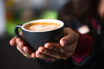 Close-up of hands holding a black cup of latte with heart-shaped latte art. Warm, cozy vibe ideal for lifestyle, coffee culture, and cafe concept.