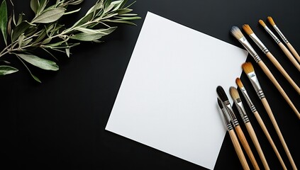 Blank canvas, art supplies on black surface.  Fresh olive branch beside a sheet of white paper, surrounded by various paintbrushes of differing sizes and shapes