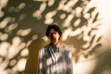 Person in white shirt standing against sunlit wall with leaf shadows