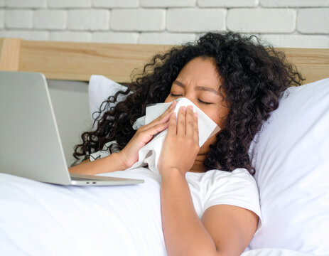 A young woman with curly hair lies in bed, looking unwell, holding a tissue to her face while a laptop is open beside her. The image suggests working while sick, or general illness and discomfort.