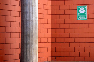 Emergency meeting point sign, in portuguese, on a red brick wall next to a cylindrical column....