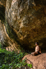 Woman meditating in a natural cave. Serene scene with rock formations and lush greenery.