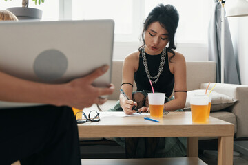 Young adult woman working in a modern workplace, focused on writing, socializing with colleagues...