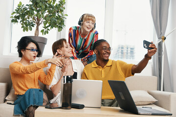 Young adult colleagues socializing with smiling man taking a selfie, diverse coworkers, casual workplace, Gen Z friends enjoying work together.
