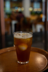 A glass of dark amber liquid, likely iced tea, with visible ice cubes, sits on a round wooden tray