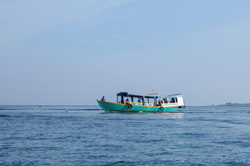 Obraz premium A vibrant turquoise and yellow passenger boat with a blue canopy sails across calm blue ocean water under a clear sky with a distant island visible on the horizon