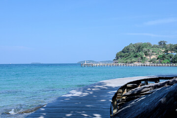 A scenic view of a curved wooden boardwalk leading towards a long pier extending into the clear turquoise ocean under a vast blue sky with a lush green island in the distance
