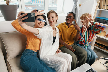 Young adult coworkers taking selfie while socializing in a bright workplace, showcasing friends from Gen Z enjoying business collaboration and work together.