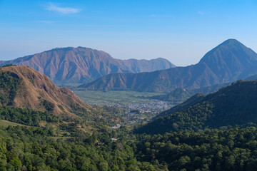 A breathtaking landscape captures Sembalun village from afar, nestled peacefully within a lush green valley surrounded by towering hills and rugged peaks under a bright blue sky in Lombok, Indonesia.