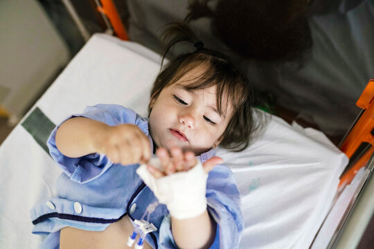 Sick toddler girl lying in hospital bed playing with bandage on hand